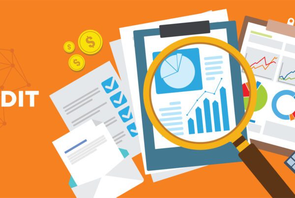 The word "audit" surrounded by papers of graphs and charts on clipboards, a calculator, and a magnifying glass